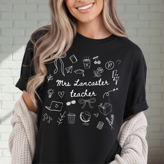 Person wearing a black doodle t-shirt with 'Mrs. Teacher' design against a white brick wall.